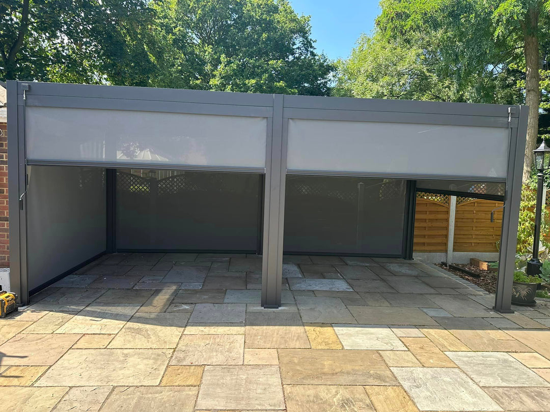 Nova Titan 6M x 3M Grey Pergola, with 6 privacy pull down blinds, on sand stone patio, with trees in back ground and blue sky.