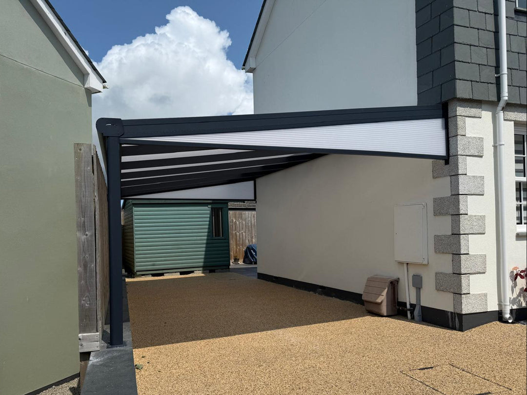 Bespoke DePonti carport installation in Cornwall, dark grey structure and opal polycarbonate roof. Installed on house andin front of a garage. with sand coloured stone driveway.