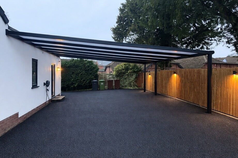 Brand new DePonti BOSCO Premium Carport installed by Midland Garden Furniture with wooden fence to side and back with new led warm white lighting and new tarmac driveway at dusk. On white house to left.
