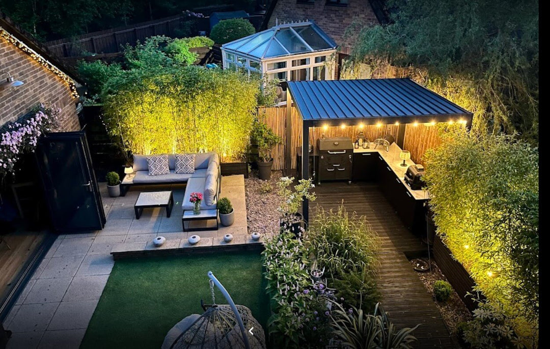 Back garden at dusk, with grey aluminium pergola, outdoor patio and garden kitchen. With nice bushes around all lit up with LED Lighting