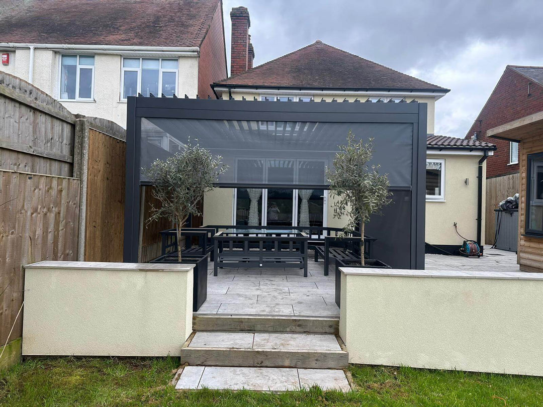Raised patio area on a cream house, with a Grey Nova Titan Aluminium Pergola with privacy screens down and 2 bushes one either side. Garden furniture in grey underneath.