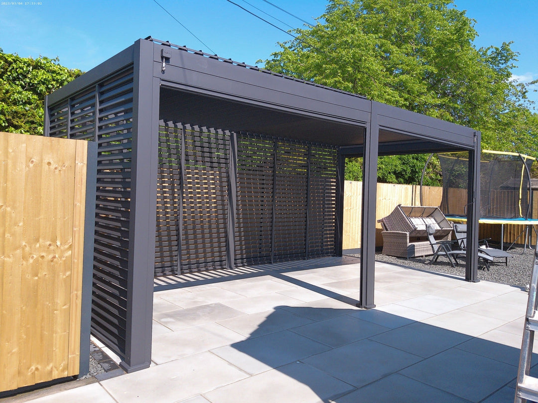 Grey Titan Pergola 6M x 3M with aluminium louvered walls at the back. Bright sunny day day against a wooden fence and big bright green trees in the background.
