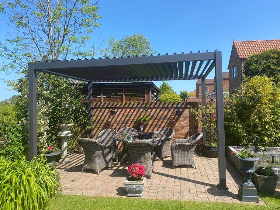 Aluminium Pergola on bright sunny day in back garden with clear blue sky and garden furniture below.