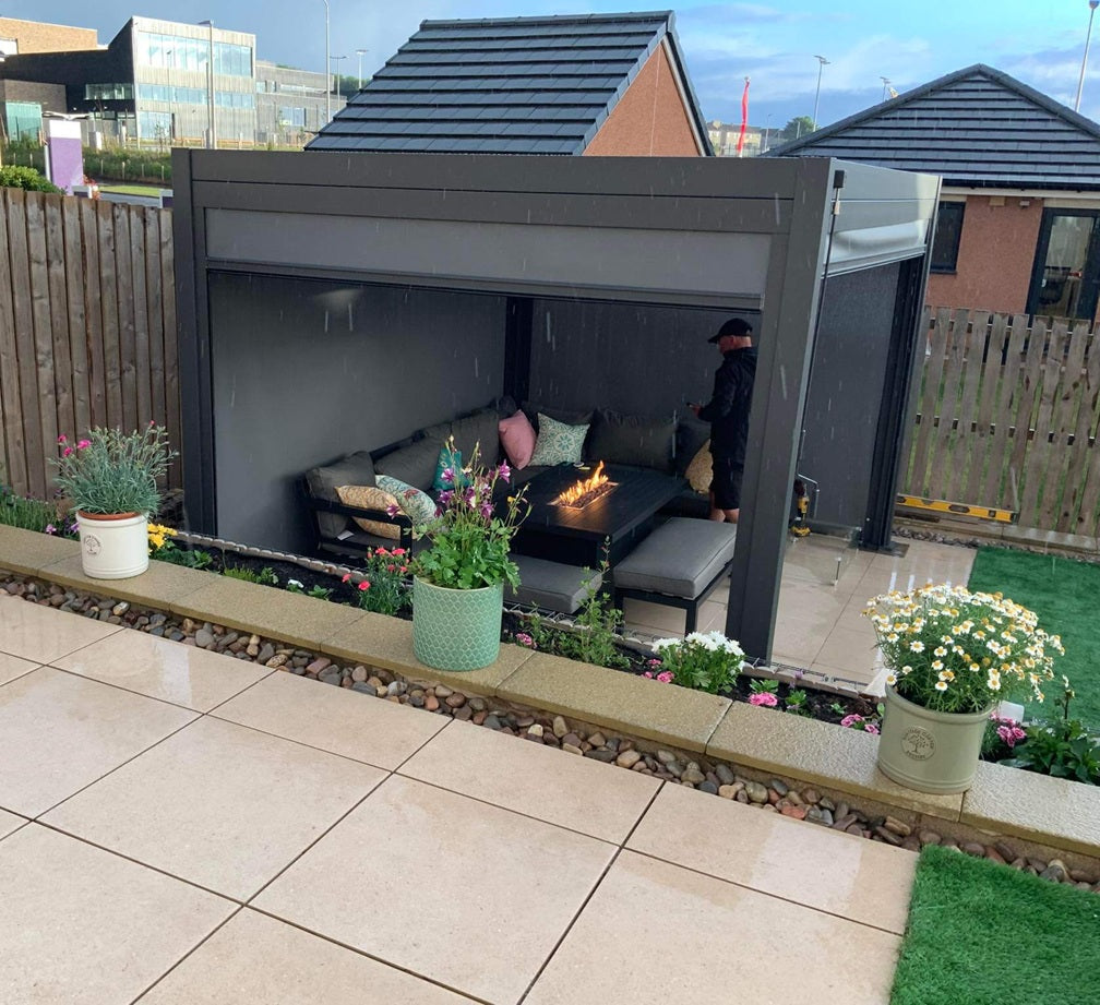 Grey Pergola in a back garden with grey sofa and chair with fire pit table with 3 potted plants in front.
