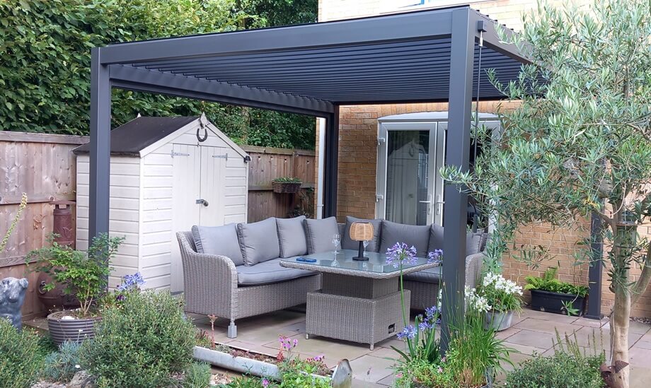 Grey Aluminium Pergola in back garden, with small tree to right and garden plants at front, with outdoor furniture underneath.