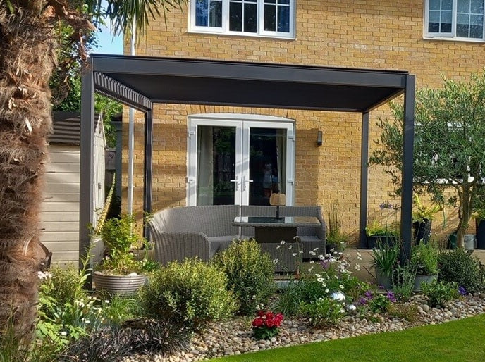 Cotswold Brick house back garden with a modern Titan Grey pergola and outdoor furniture under, with mature flowers in front and palm tree to left. Beautiful cottage style garden.