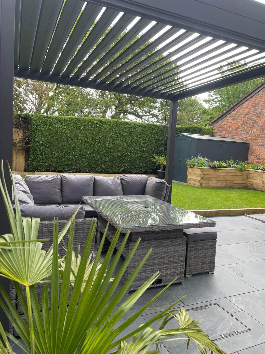Grey Nova Titan pergola installed in back garden of new brick house, on grey tiles floor. Green grass in front and cloudy blue sky.