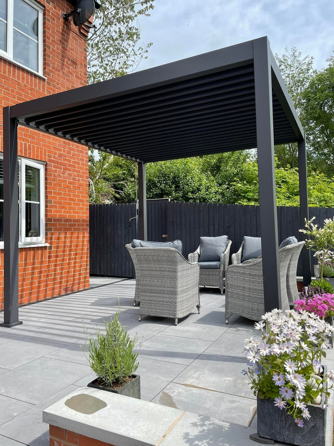 Modern Grey Pergola on Porcelain slabs, in back garden beautiful flowers to the right and garden furniture underneath