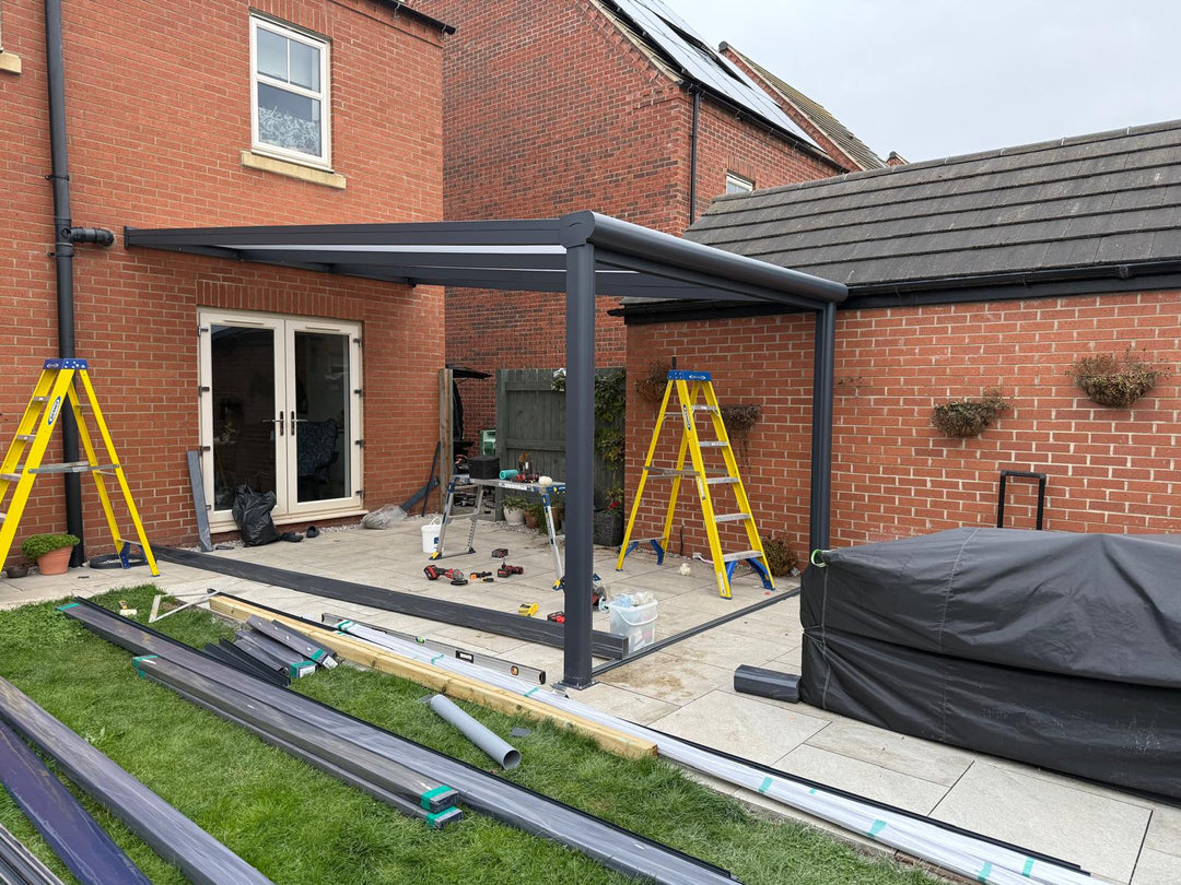 A modern Deponti veranda under construction by Midland Garden Furniture, featuring a sleek anthracite metal frame with a translucent opal white roof. 
The structure is installed on a cream titled patio, with ladders and tools being used for installation. The veranda is awaiting glass doors to be fitted.