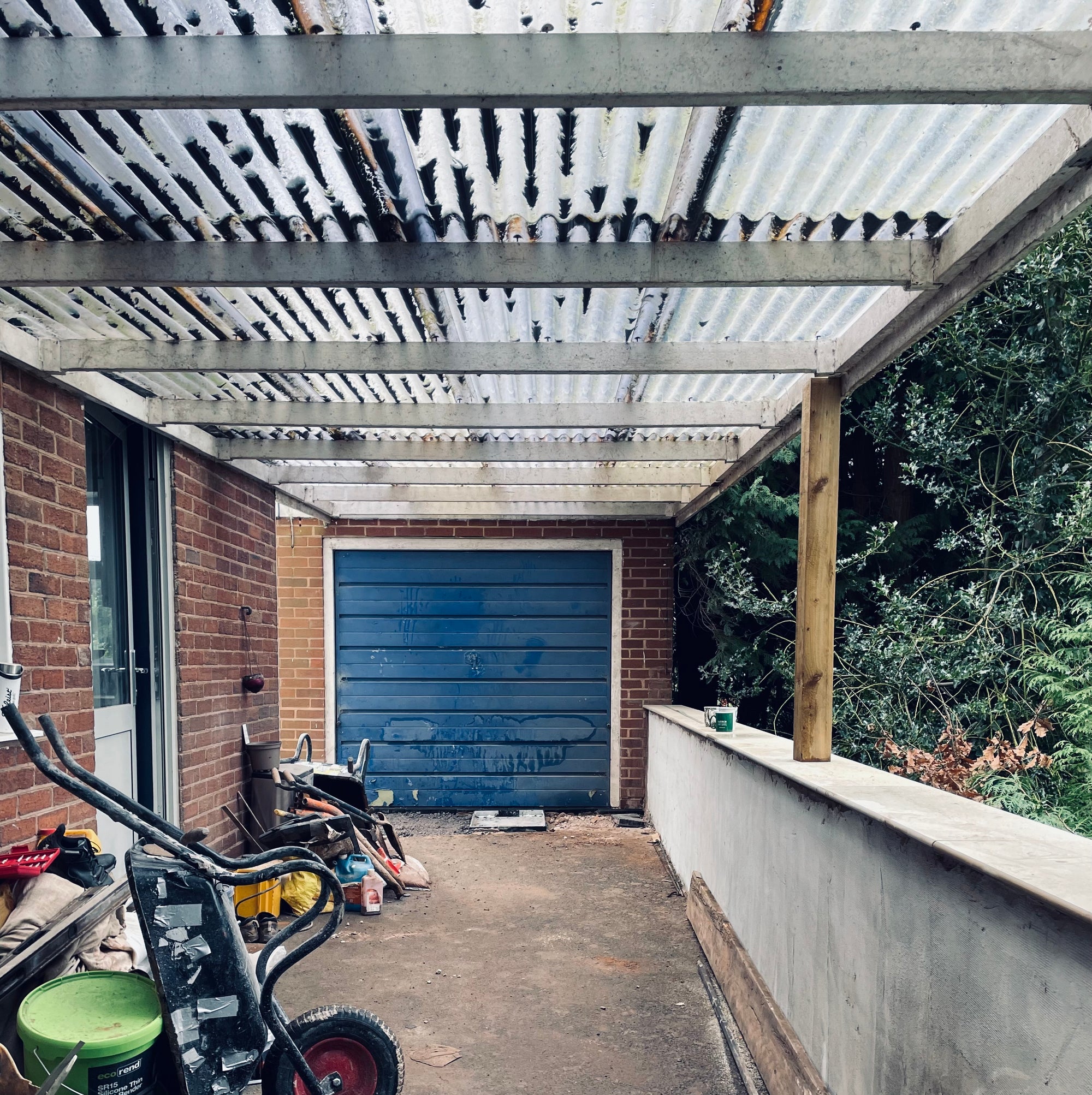 Covered outdoor area old carport with moss on the translucent roof, looks old and derelict. With a blue garage door, brick wall, and greenery and shrubs to the right. The garage is awaiting to be upgraded to a Midland Garden Furniture DePonti carport. 