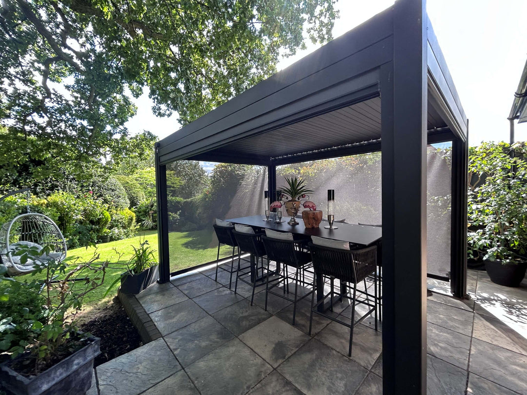 A modern black outdoor patio and NOVA TITAN Pergola from Midland Garden Furniture with a dining table and six bar stools underneath, set on a stone tile patio in a lush green backyard. Aylesbury Oxfordshire
