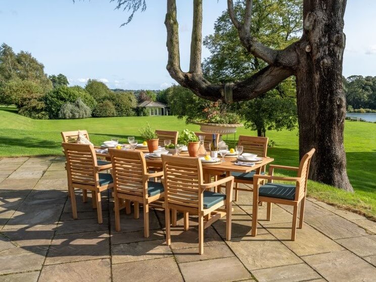 Hanover Teak Outdoor dining furniture with wooden table and chairs on a patio, surrounded by greenery.