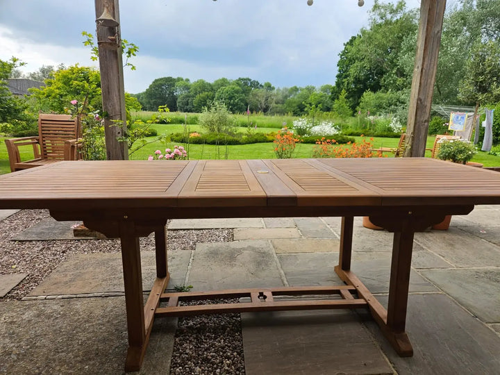 Hanover Teak  Wooden garden table on a patio with a scenic background of trees and flowers.
