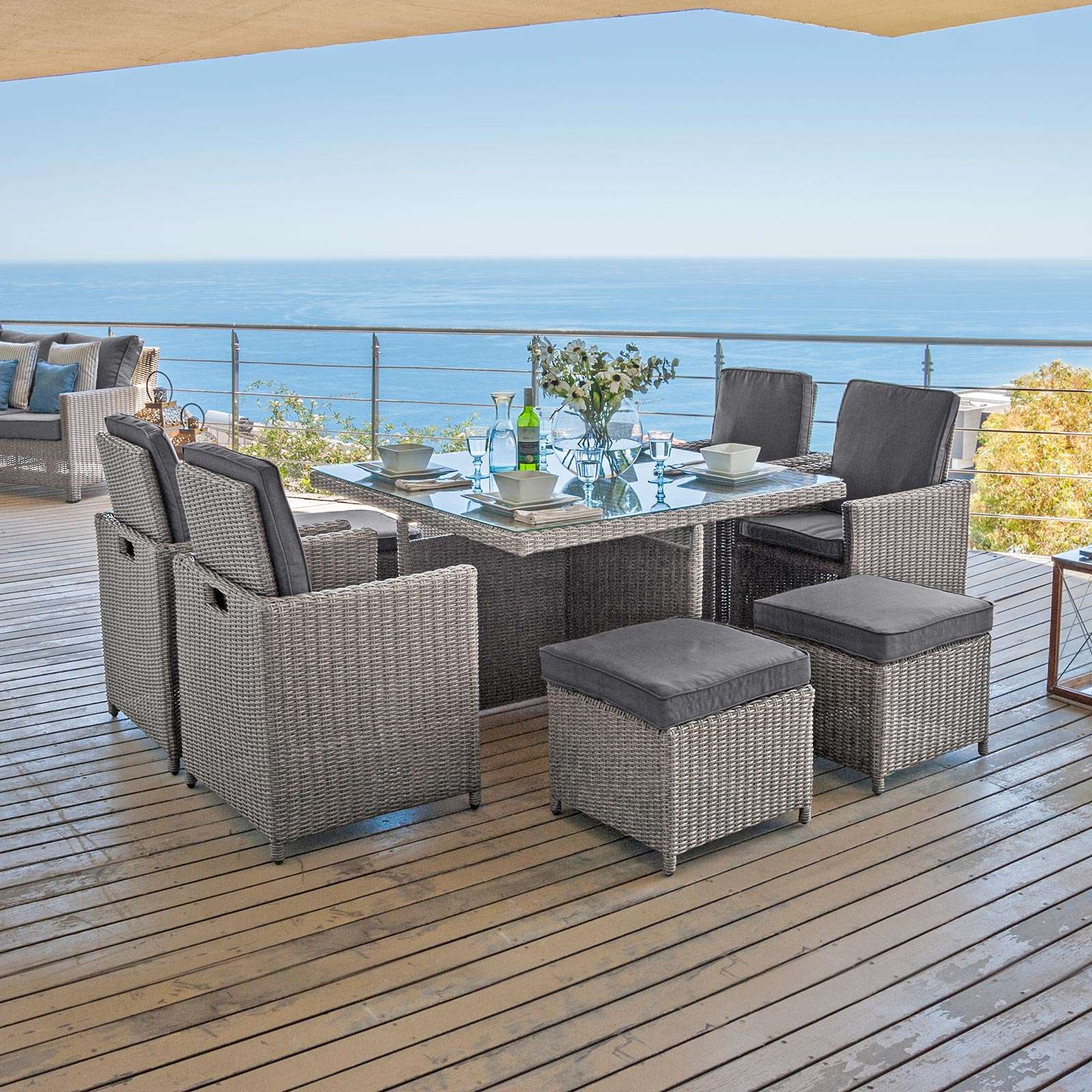 Light grey with dark grey cushions cube garden furniture with glass top table. On a wooden decking overlooking the sea. 