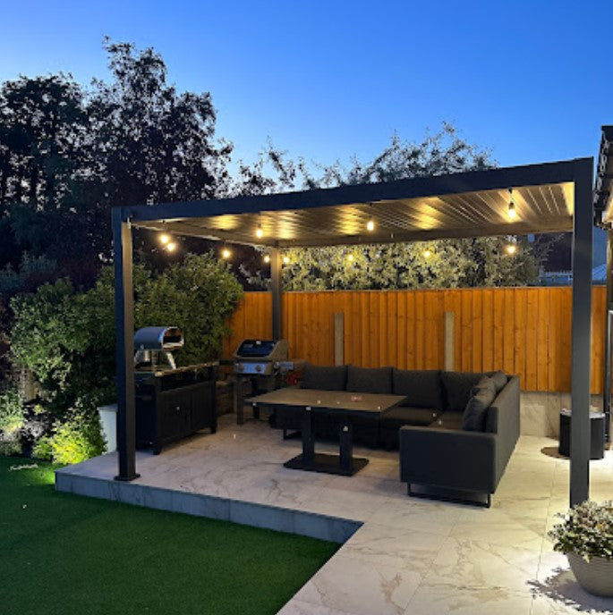 Nova Titan Grey Pergola at dusk with LED lights on marble tiles and dark grey outdoor sofa and outdoor kitchen.