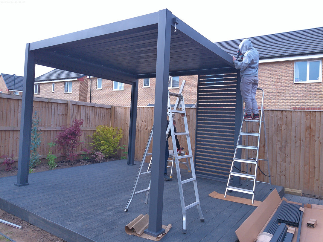 Anthracite grey Nova Titan Pergola being installed by person on ladders on grey decking. Wooden fence around the back.