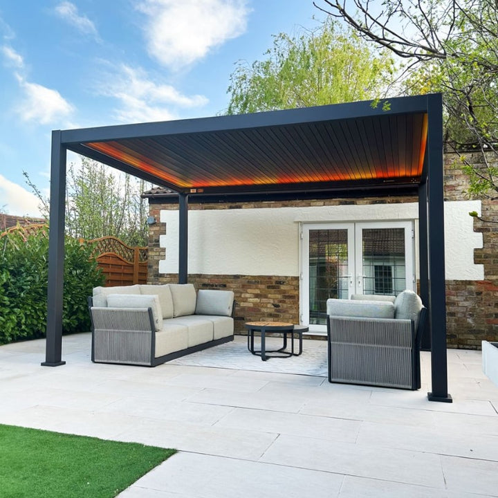 Nova Titan Plus Pergola Grey on white slabs in garden in front of garage. Orange coloured LED lighting under the Pergola roof, and a modern light grey roped outdoor sofa.