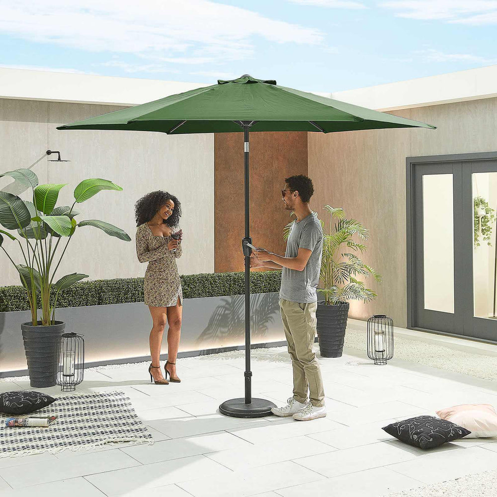 Two people standing under a large green NOVA Outdoor living patio parasol in an outdoor setting.