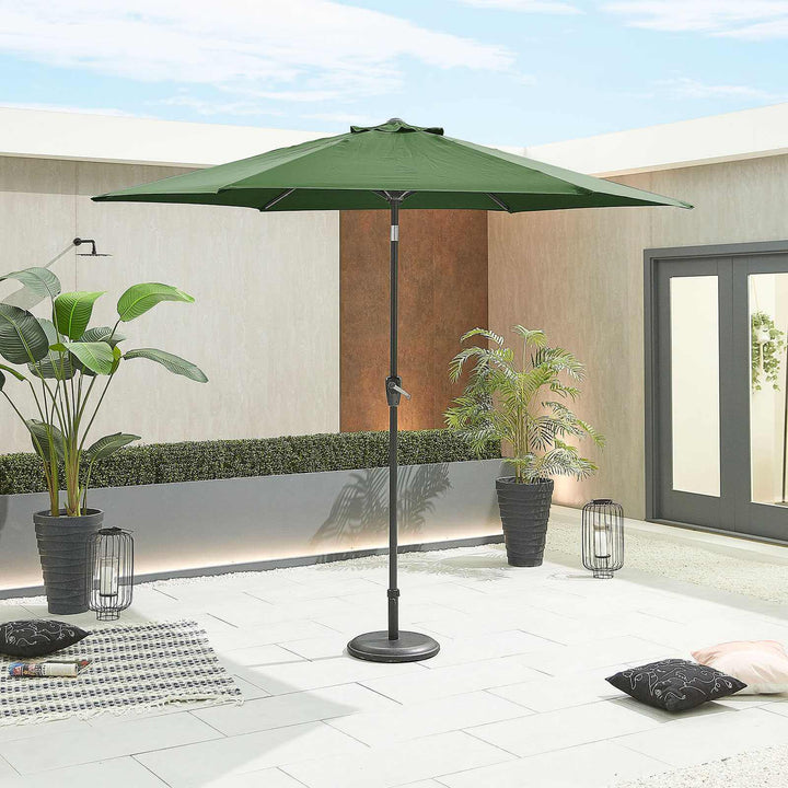 Green patio NOVA Outdoor living parasol on a stand in an outdoor setting with plants and a building in the background.