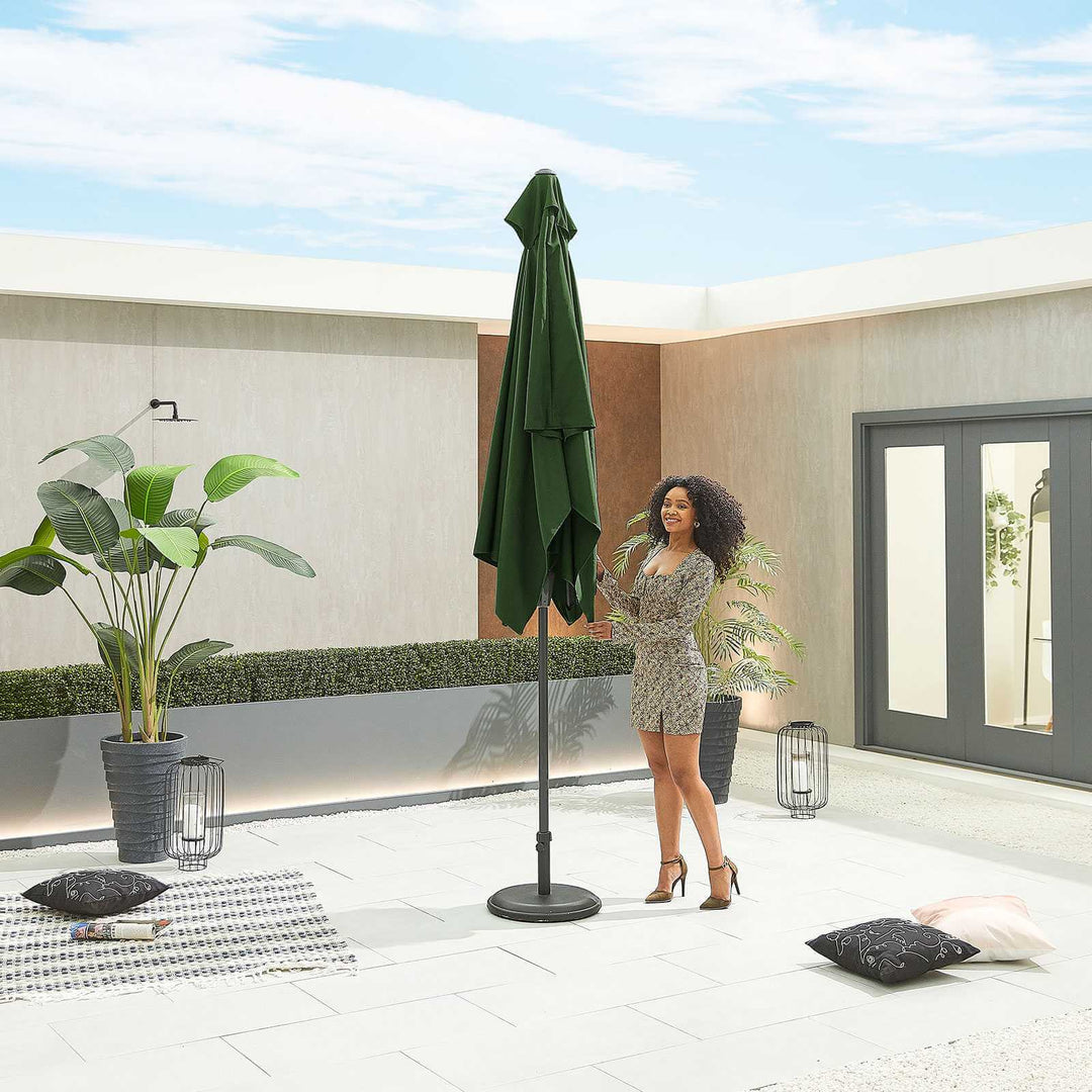 Woman standing next to a green Nova Outdoor Living Parasol on a modern patio with plants and a door in the background.