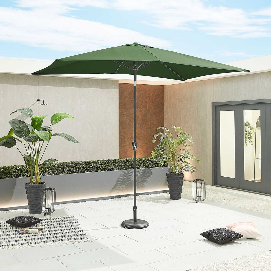 Green patio Nova Outdoor Living Parasol on a stand in an outdoor setting with plants and cushions.