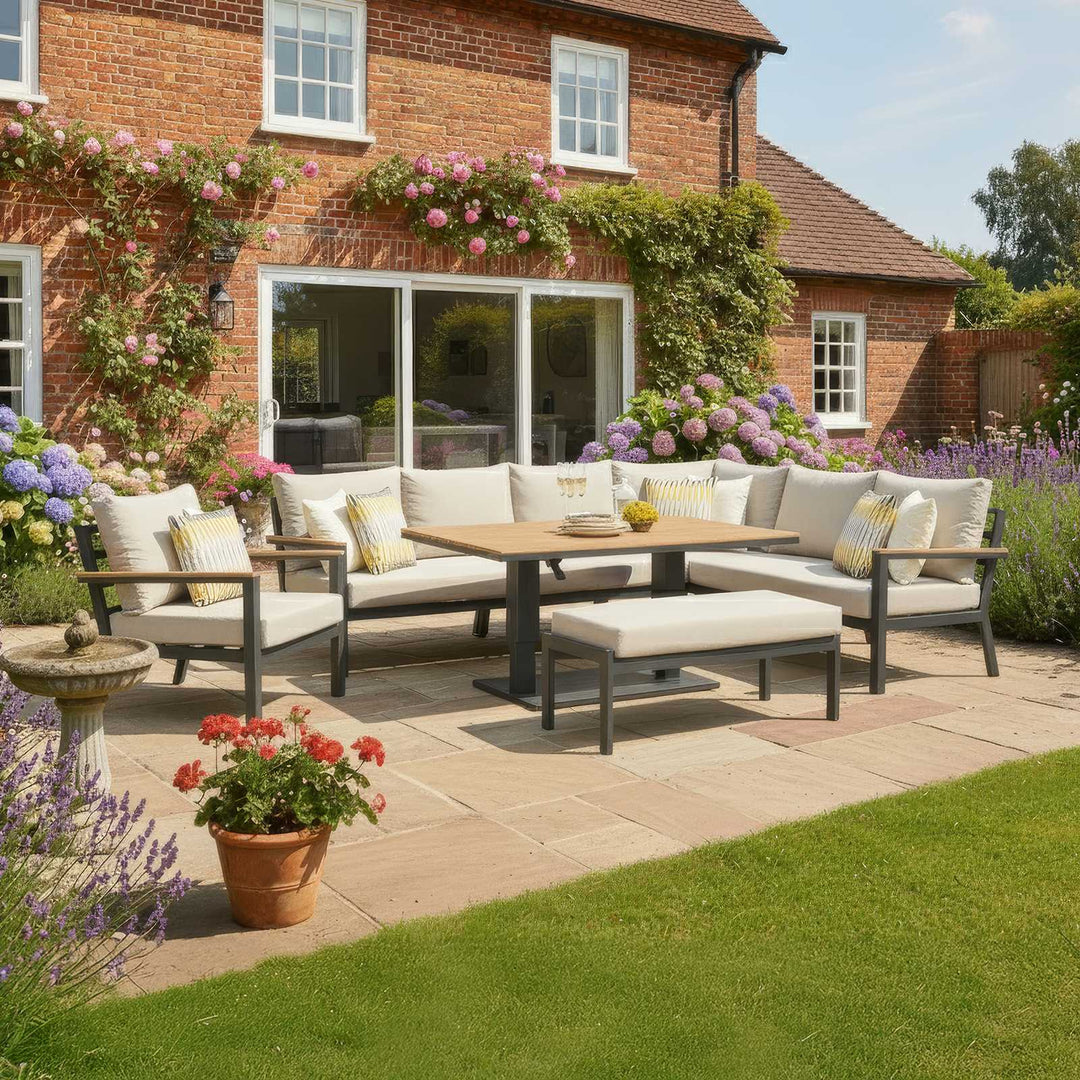 Outdoor Nova Heidi patio set with light grey/ beige cushions with darg grey aluminium frame in front of a brick house with floral decorations on stone slab patio area. Right Hand L Shaped sofa