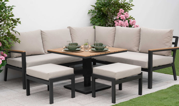 Outdoor Nova Heidi patio set with light grey/ beige cushions with dark grey aluminium frame in front of a plain white wall with green grass to the front and plants either side. Square with 2 small foot stools.