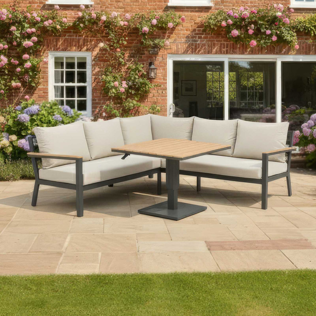 Outdoor Nova Heidi patio set with light grey/ beige cushions with dark grey aluminium frame in front of a brick house with floral decorations on stone slab patio area. Rose flowers trailing up the house walls.