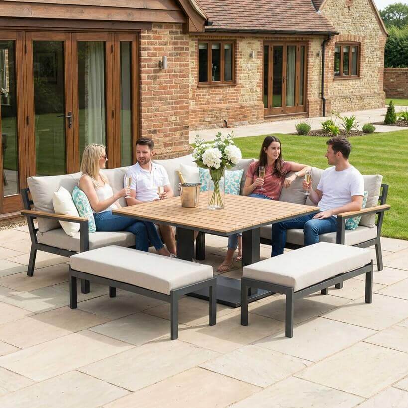 Nova Heidi outdoor living furniture set Midland Garden Furniture with people enjoying in front of a Cotswold house with plants at the back. corner sofa set with two benches and rising table. 
