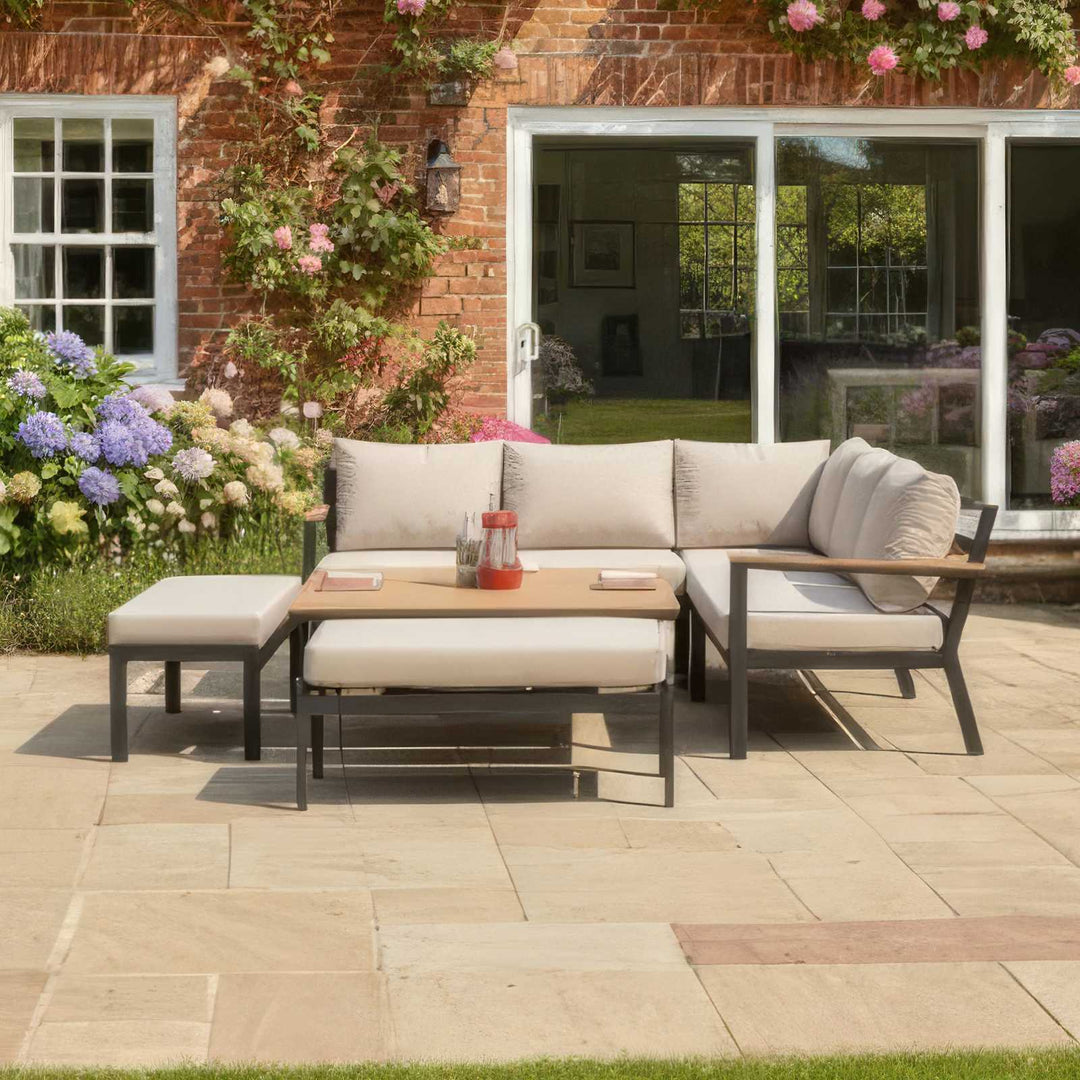 Outdoor Nova Heidi patio set with light grey/ beige cushions with darg grey aluminium frame in front of a brick house with floral decorations on stone slab patio area.