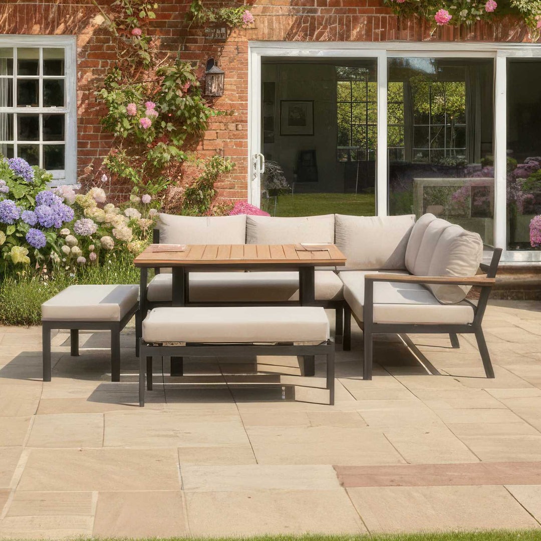 Outdoor Nova Heidi patio set with light grey/ beige cushions with darg grey aluminium frame in front of a brick house with floral decorations on stone slab patio area. With Bench and stool
