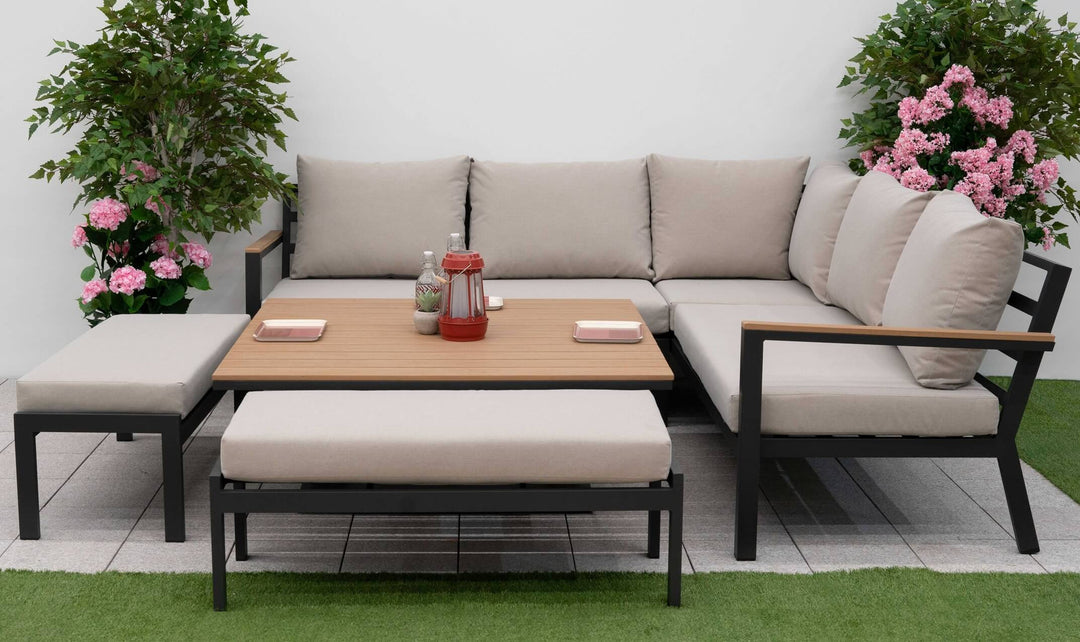Outdoor Nova Heidi patio set with light grey/ beige cushions with dark grey aluminium frame in front of a plain white wall with green grass to the front and green and pink plants either side.