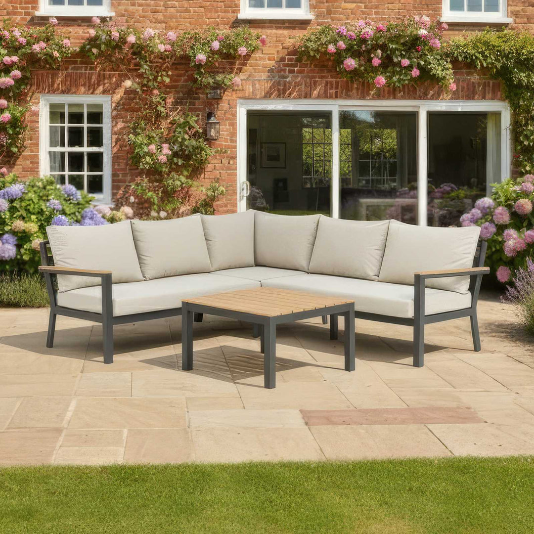 Outdoor Nova Heidi patio set with light grey/ beige cushions with dark grey aluminium frame in front of a brick house with floral decorations on stone slab patio area. Green well cut lawn to front.