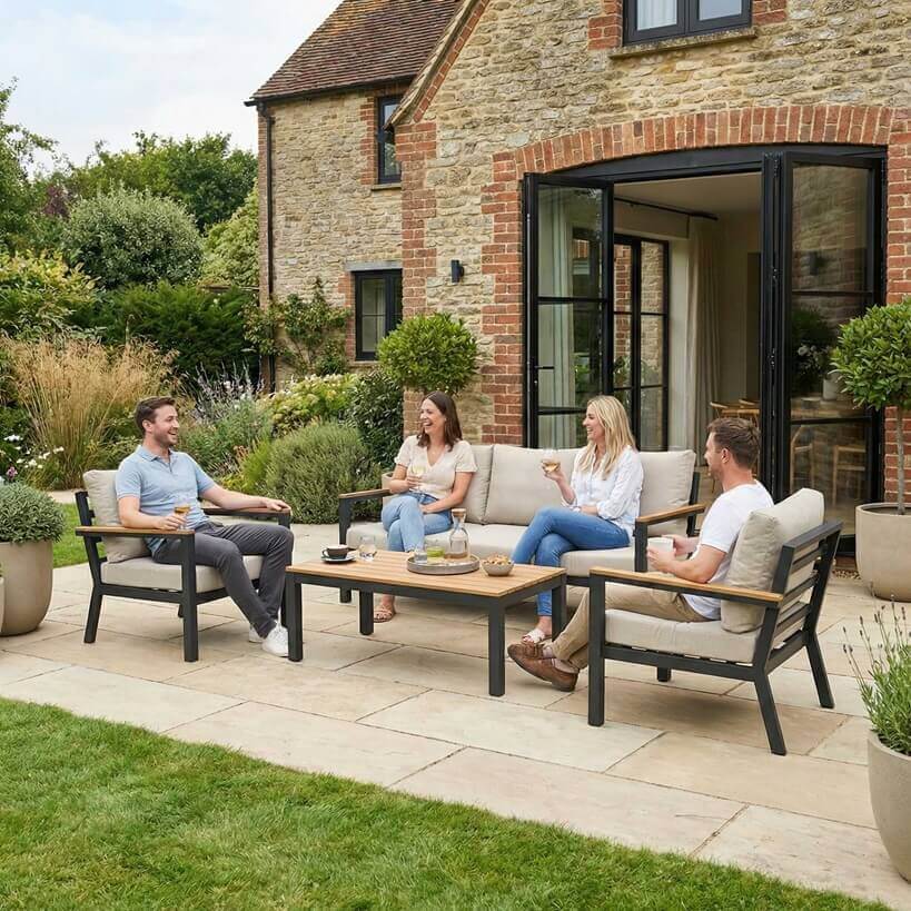 Nova Heidi outdoor living furniture set Midland Garden Furniture with people enjoying in front of a Cotswold house with plants at the back. sofa set with 3 seater sofa and 2 arm chairs and coffee table.