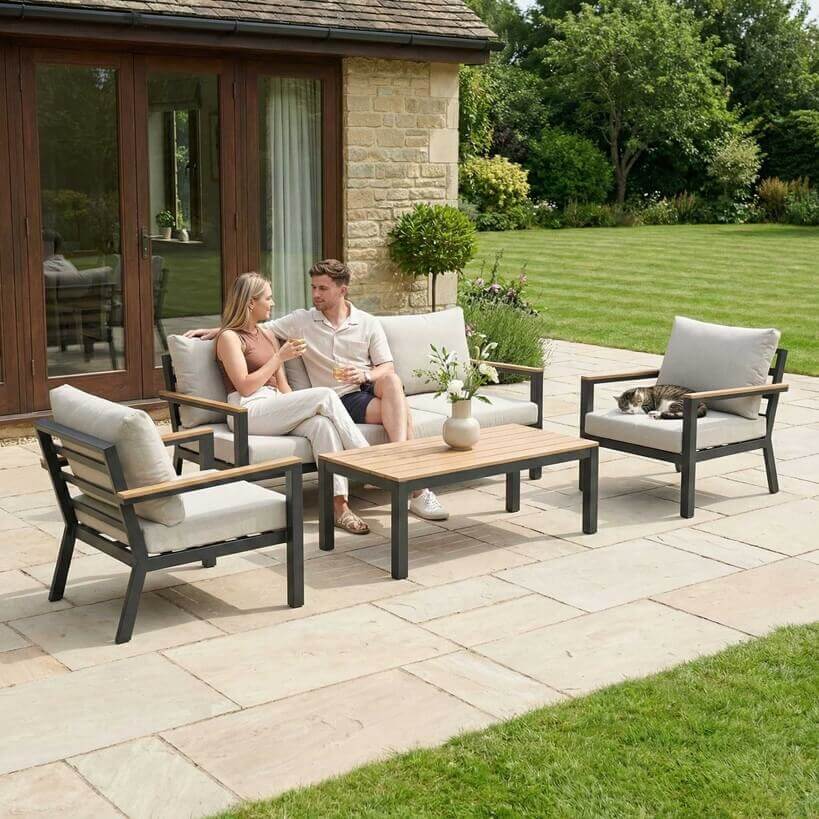 Nova Heidi outdoor living furniture set Midland Garden Furniture with people enjoying in front of a Cotswold house with plants at the back. sofa set with arm chairs and rising table. 