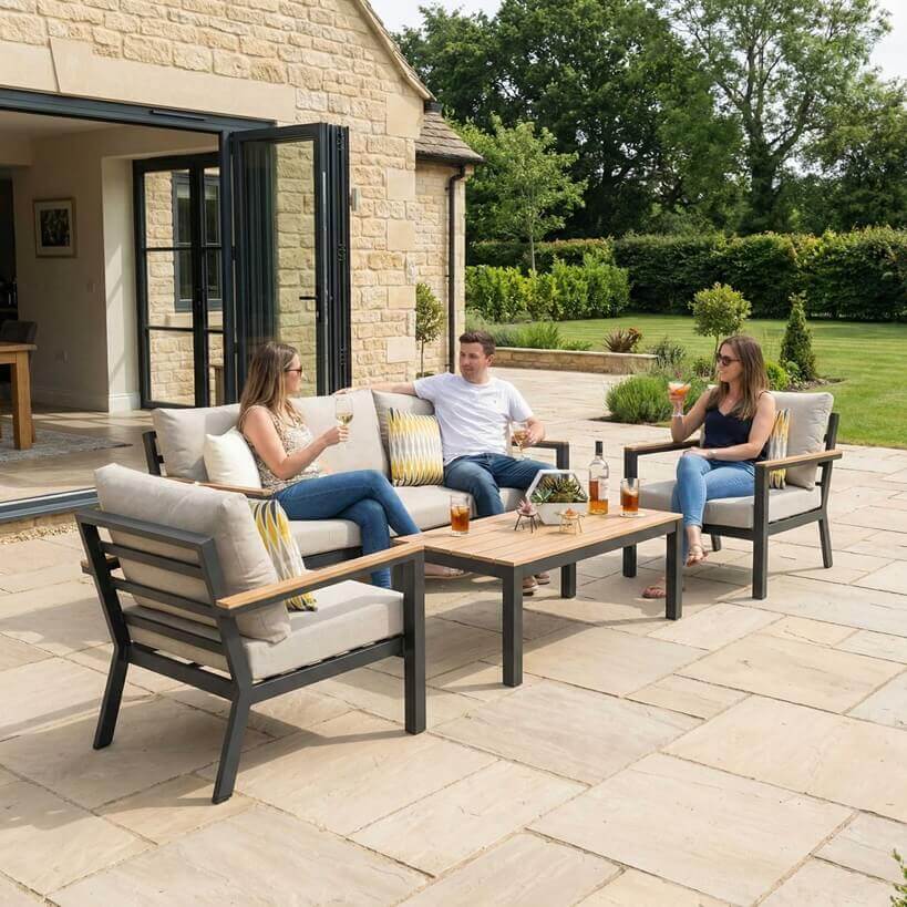 Nova Heidi outdoor living furniture set Midland Garden Furniture with people enjoying in front of a Cotswold house with plants at the back. sofa set with arm chairs and rising table. 