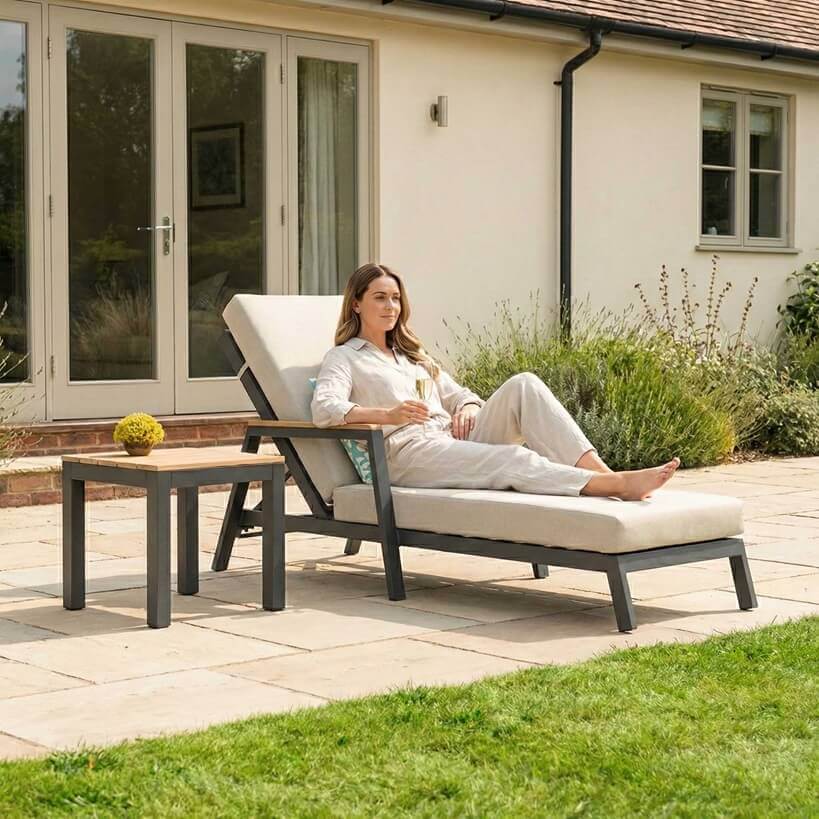 Nova Heidi outdoor living furniture set Midland Garden Furniture with lady enjoying in front of a Cotswold house with plants at the back. Sun lounger and drinks table.