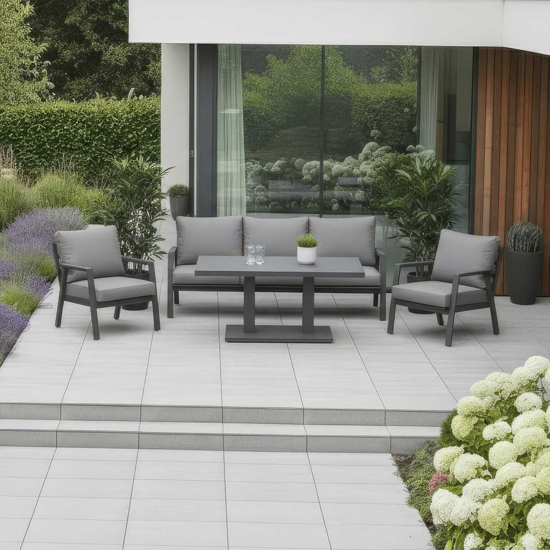 Nova Phoenix Outdoor patio set with grey sofa, chairs, and rising table on a tiled patio. In front of modern House with glass windows and flowers and shrubs to the side and back.