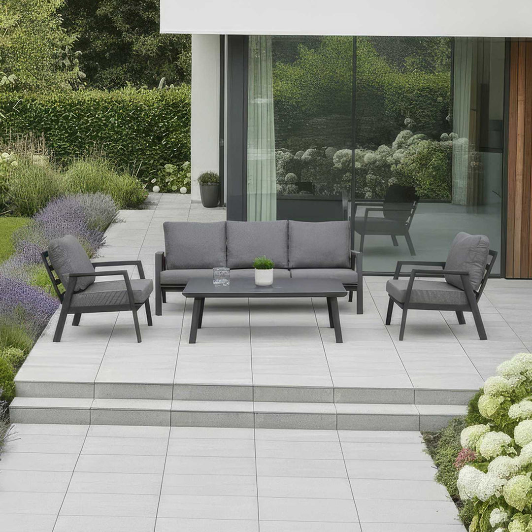 Nova Phoenix Outdoor patio set with grey sofa, chairs, and coffee  table on a tiled patio. In front of modern House with glass windows and flowers and shrubs to the side and back.