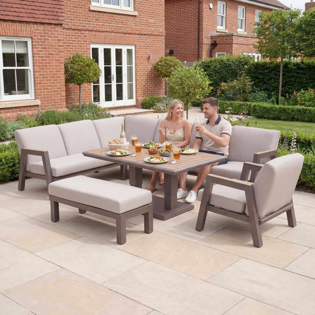 Two people sitting at a patio table with outdoor furniture in a garden setting. Nova Hunter Sofa Set Midland Garden Furniture. set in a new modern garden, with trees and shrubs at back of big house.