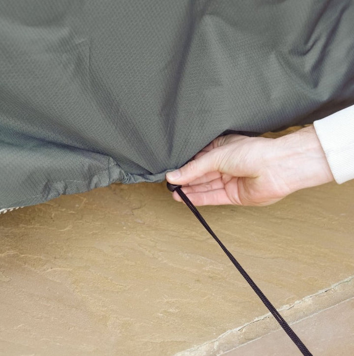 Person tightening a pullcord to secure a grey protex garden furniture cover over a garden furniture set, over sandstone paving