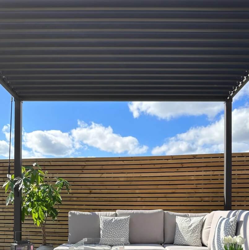 Outdoor patio with a Nova Luna pergola, wooden fence, and outdoor furniture under a clear blue sky.