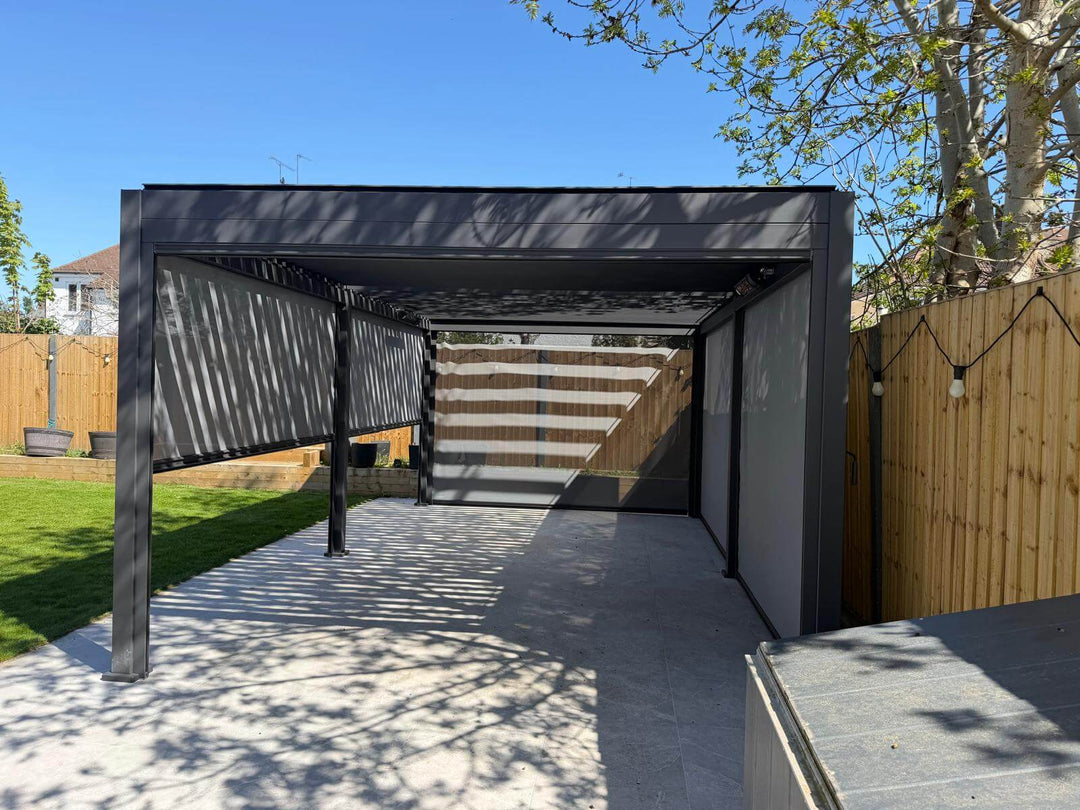 6M x 3M Nova Titan Aluminium Pergola installed in Redhill, Surrey by Midland Garden Furniture on new patio slabs with grass to side and tress behind on bright sunny morning. with 6 Pergola privacy screens all pulled down 