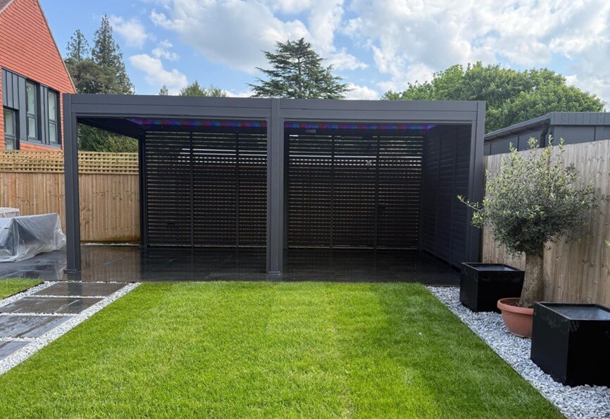 Titan Pergola 6M x 3M with multicolour LED lighting and aluminium louvered walls to the back and green grass tot he front with garden fence at the back