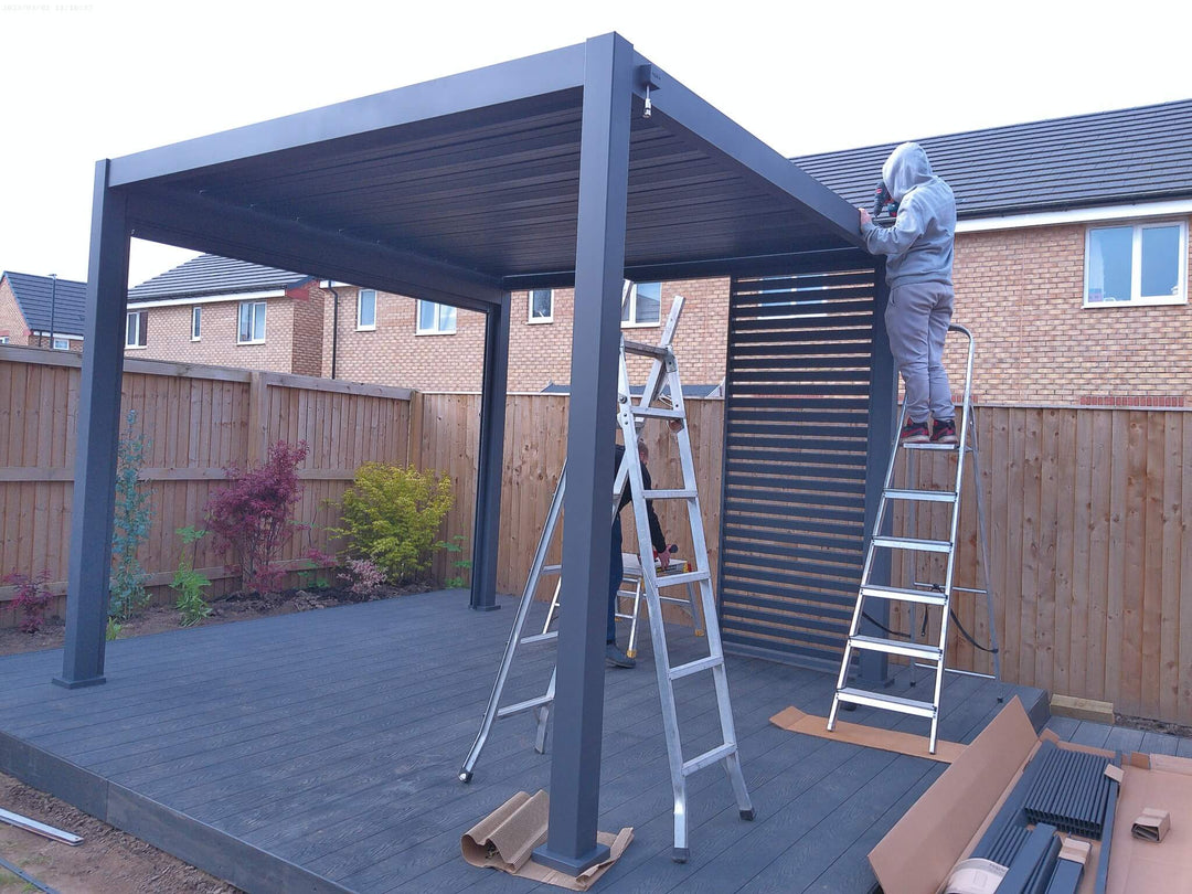 Grey Nova Titan Pergola being installed on grey decking with garden fence to the back with houses in background on new build estate. Work up a ladder putting on the roof.