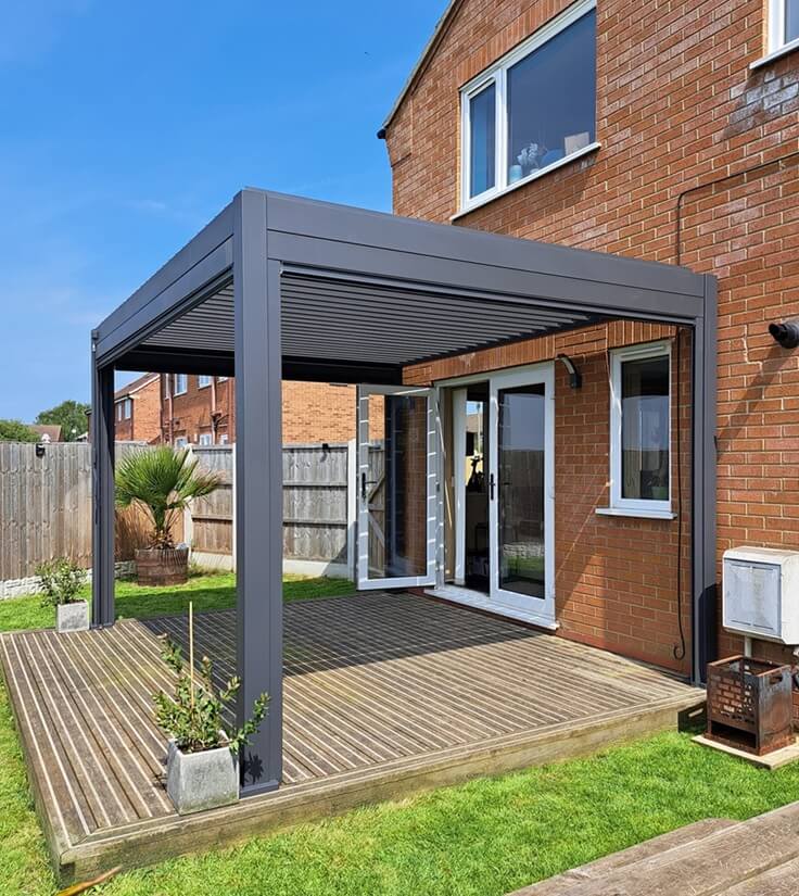 Wooden decking decking in back garden next to house, with an Aluminium pergola next to a brick house on a sunny day.