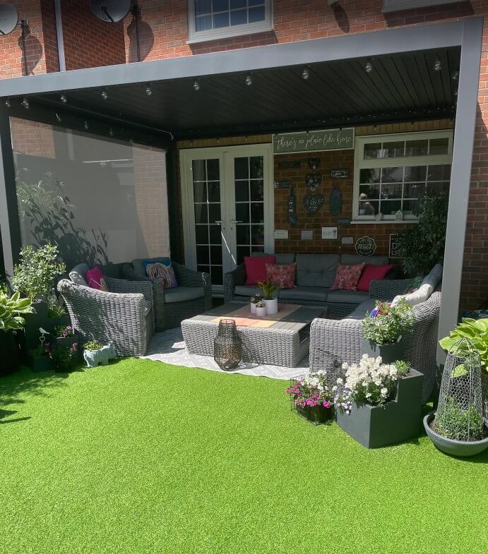 Outdoor back garden patio area with rattan grey garden furniture underneath and plants in front of a house with green artificial grass to the front.