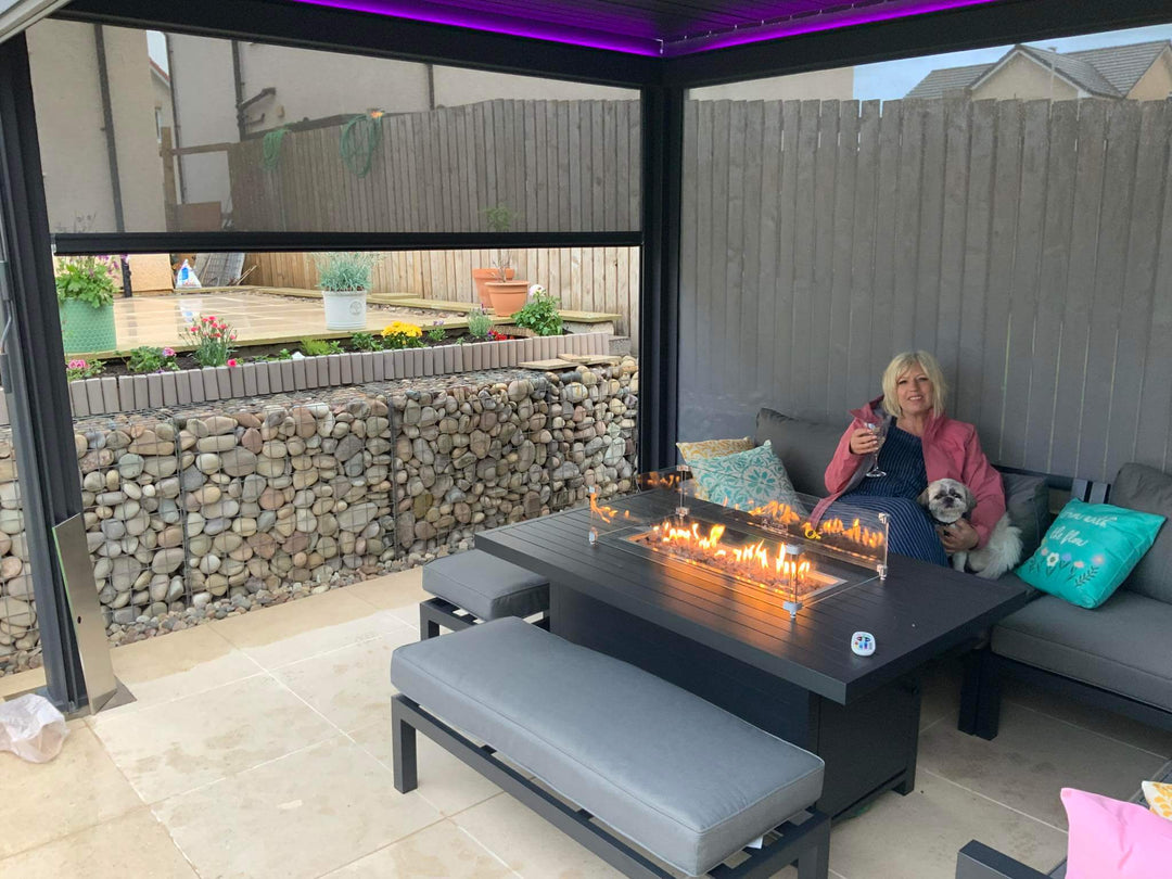 Woman sitting inside a Pergola on a patio with a fire pit table and outdoor furniture. With her small dog and glass of wine. 