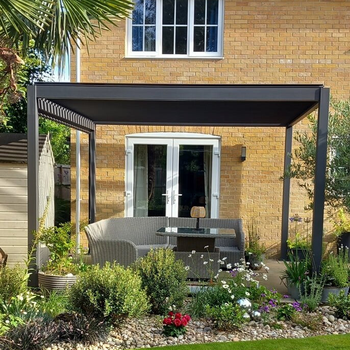 Cotswold Brick house back garden with a modern Titan Grey pergola and outdoor furniture under, with mature flowers in front and palm tree to left.