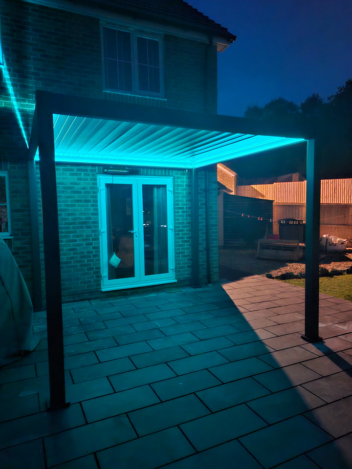 Nova Titan Plus Pergola at night with light blue LED lighting under the roof. against a house with French doors into Pergola.